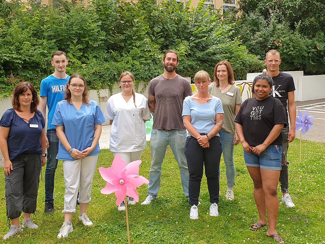 Gruppe von neun Personen steht auf einer Wiese vor einem Hintergrund aus Büschen und Bäumen, im Vordergrund zwei große bunte Windräder aus Kunststoff